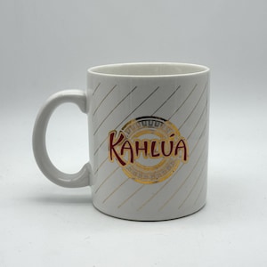 May include: White ceramic mug with the Kahlúa logo in a gold and red circular design. The mug has a gray handle and diagonal gray and gold stripes. A coffee mug for enjoying your favorite beverage.