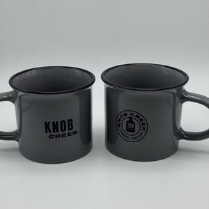 May include: Two gray ceramic mugs with black rims and handles. The left mug features the black text "KNOB CREEK." The right mug displays a circular black logo with the text "KNOB CREEK" and "CLERMONT, KENTUCKY" surrounding a "100" graphic.