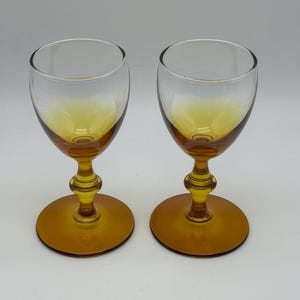 May include: Two amber-colored wine glasses. The bowls are clear, transitioning to amber at the base. They have a decorative stem and a round base. The glasses are set against a plain white background.