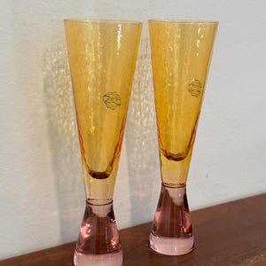 May include: Two tall, amber-colored champagne flutes with textured glass. The stems are a soft pink, and each glass has a small, circular label near the top. The glasses are displayed on a dark wooden surface.
