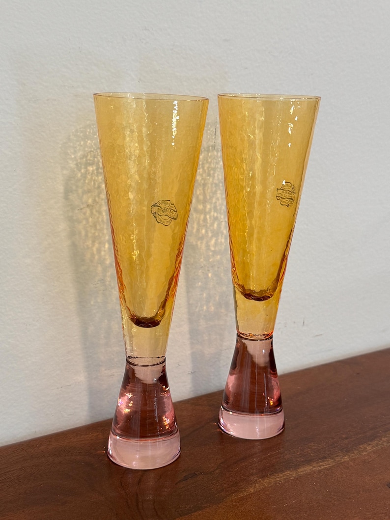 May include: Two tall, amber-colored champagne flutes with textured glass. The stems are a soft pink, and each glass has a small, circular label near the top. The glasses are displayed on a dark wooden surface.