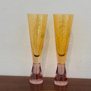 May include: Two champagne flutes with a textured, amber-colored bowl and a pink base. The flutes are displayed on a wooden surface against a white wall. The stemware has a classic, elegant design.