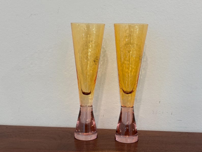 May include: Two champagne flutes with a textured, amber-colored bowl and a pink base. The flutes are displayed on a wooden surface against a white wall. The stemware has a classic, elegant design.