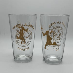 May include: Two clear glass pint glasses featuring brown printed graphics. Each glass displays the text Vanilla Java Porter, Atwater Brewery, Born in Detroit, Raised Everywhere, and an illustration of a person in a tuxedo holding a tray.