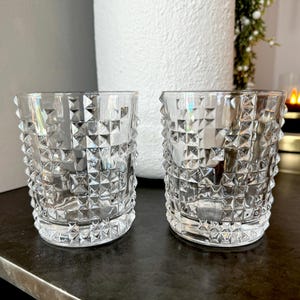 May include: Two clear, faceted drinking glasses. The glasses have a low, rounded shape and are covered in a repeating diamond pattern. The glasses are sitting on a dark surface.