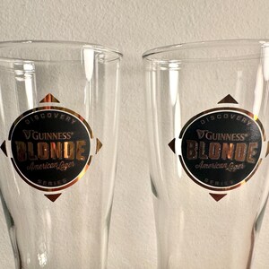 Puede incluir: Dos vasos de pinta de vidrio transparente con un logotipo dorado y negro de Guinness Blonde American Lager. El logotipo dice "Discovery Series" y presenta una estrella dorada.