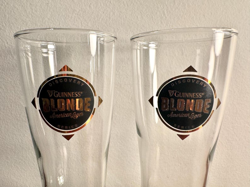 Puede incluir: Dos vasos de pinta de vidrio transparente con un logotipo dorado y negro de Guinness Blonde American Lager. El logotipo dice "Discovery Series" y presenta una estrella dorada.