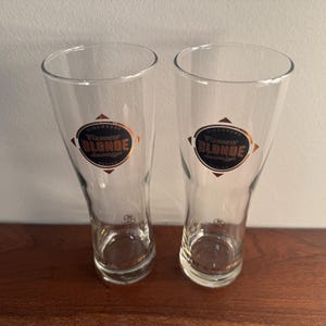 Puede incluir: Dos vasos de pinta de vidrio transparente con una etiqueta dorada y negra que dice "Guinness Blonde Lager".