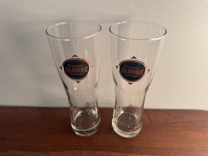 Puede incluir: Dos vasos de pinta de vidrio transparente con una etiqueta dorada y negra que dice "Guinness Blonde Lager".