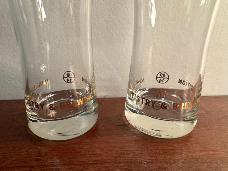 Puede incluir: Dos vasos de beber de vidrio transparente con letras doradas que dicen "Brewery & Distillery" y un c&iacute;rculo dorado con el n&uacute;mero "60" en su interior.