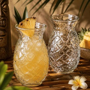 May include: Two clear glass tumblers shaped like pineapples with a textured diamond pattern. One glass is filled with a yellow liquid, ice cubes, and a fresh pineapple slice garnish. The other glass is empty and sits next to a white flower.