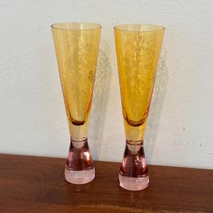 May include: Two tall, elegant champagne flutes. The glasses have a textured, golden-yellow bowl that tapers to a narrow stem and a pink base. The flutes are displayed on a wooden surface against a white wall.