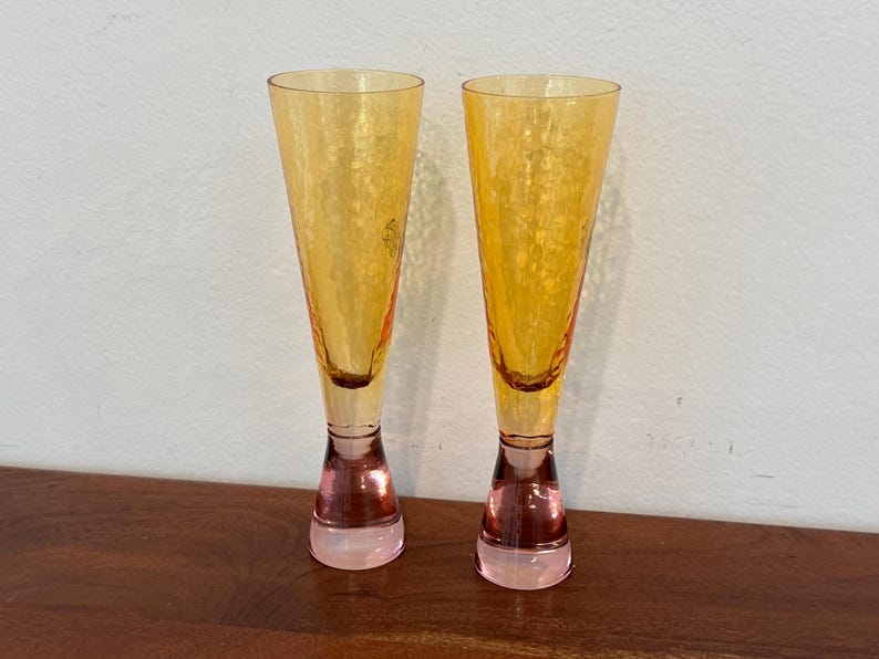 May include: Two tall, elegant champagne flutes. The glasses have a textured, golden-yellow bowl that tapers to a narrow stem and a pink base. The flutes are displayed on a wooden surface against a white wall.