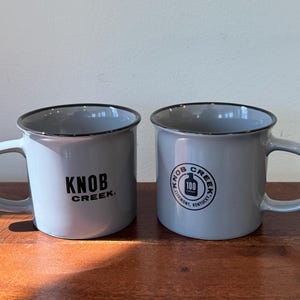 May include: Two gray ceramic mugs with black handles and black lettering. The mug on the left says "Knob Creek." The mug on the right says "Knob Creek Clermont, Kentucky."