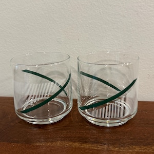 May include: Two clear drinking glasses with a green diagonal stripe design. The glasses have vertical white lines. The glasses are sitting on a wooden surface.