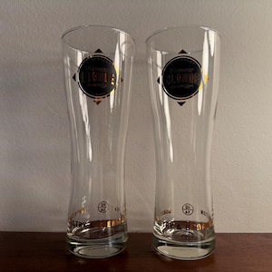 Puede incluir: Dos vasos de cerveza de vidrio transparente con letras doradas. Los vasos son altos y delgados con una base redondeada. El texto en los vasos dice "St. Lawrence's Blonde" y "St. Lawrence's Original".