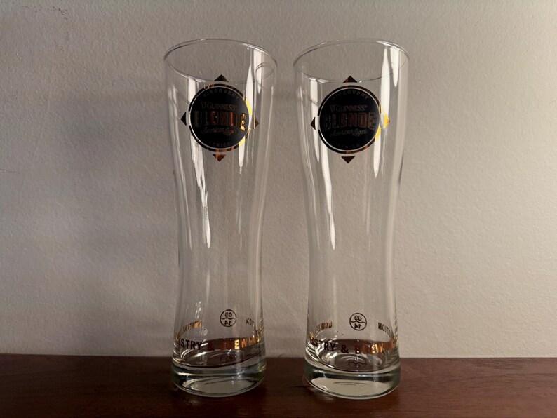 Puede incluir: Dos vasos de cerveza de vidrio transparente con letras doradas. Los vasos son altos y delgados con una base redondeada. El texto en los vasos dice "St. Lawrence's Blonde" y "St. Lawrence's Original".
