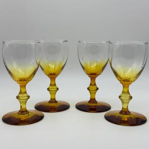 May include: Four amber-colored wine glasses with clear bowls and ornate stems. The glasses have a classic design, with a rounded bowl and a detailed stem. The bases are circular and the glasses are arranged on a white surface.