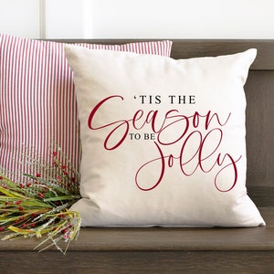 May include: A white cushion with red text that reads 'Tis the season to be jolly' on a wooden bench with a red and white striped cushion and a sprig of greenery with red berries.