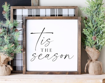 May include: A white sign with black text that reads "'Tis the season" in a cursive font. The sign is framed in a dark wood frame and is sitting on a wooden shelf with two small artificial pine trees and a white ceramic coffee canister.