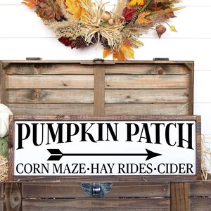 May include: A white wooden sign with black text that reads "Pumpkin Patch" with an arrow pointing to the right. Below the text, it reads "Corn Maze - Hay Rides - Cider". The sign is sitting on top of a wooden crate with a fall wreath hanging above it.