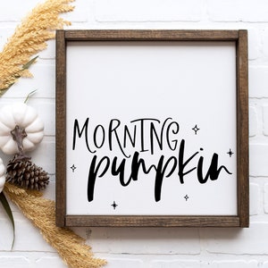 May include: A wooden framed sign with a white background and black lettering that reads "Morning Pumpkin". The sign is decorated with small stars. The sign is sitting on a white brick wall with white pumpkins and dried grasses.