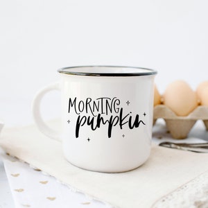 May include: White ceramic mug with a black rim and the text "MORNING pumpkin" in black script. The mug is sitting on a white napkin with gold hearts.