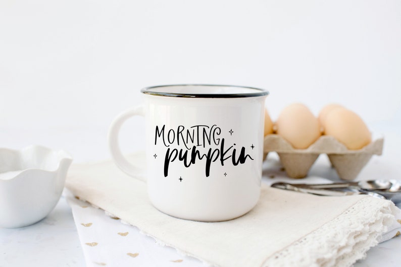 May include: White ceramic mug with a black rim and the text "MORNING pumpkin" in black script. The mug is sitting on a white napkin with gold hearts.