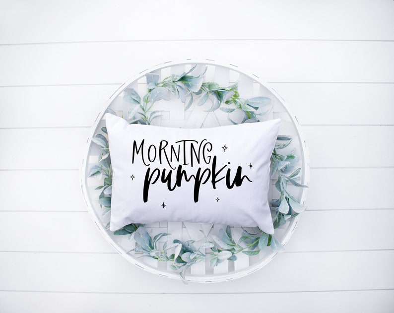 May include: White pillow with black text that reads "Morning Pumpkin" in a decorative font. The pillow is sitting in a white round basket with green leaves.