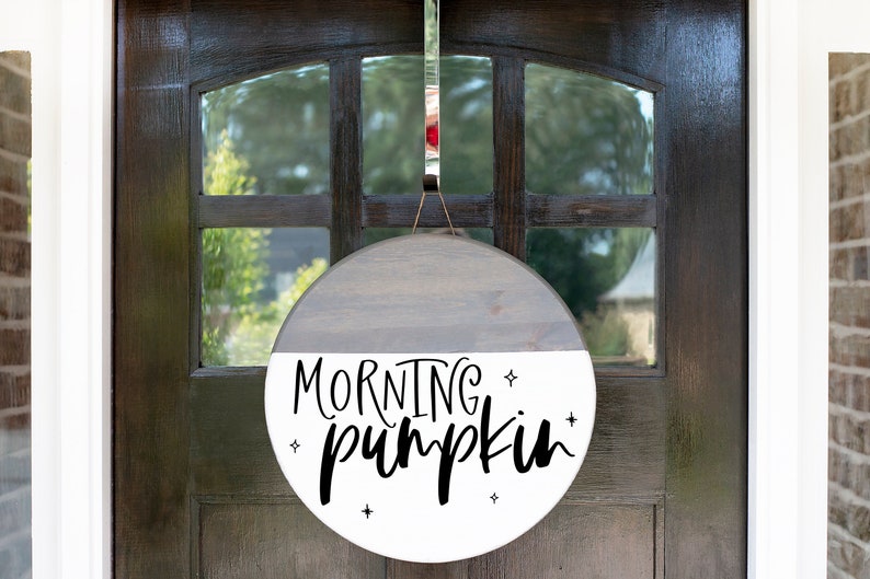 May include: A round wooden door hanger with a gray and white painted finish. The text "Morning pumpkin" is written in black script on the white half of the hanger.