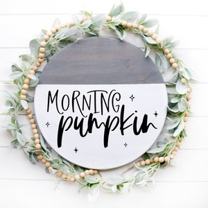 May include: A round wooden sign with a gray and white painted finish. The sign has the text "MORNING pumpkin" written in black script with small stars around the text.