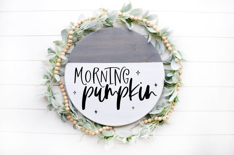 May include: A round wooden sign with a gray and white painted finish. The sign has the text "MORNING pumpkin" written in black script with small stars around the text.