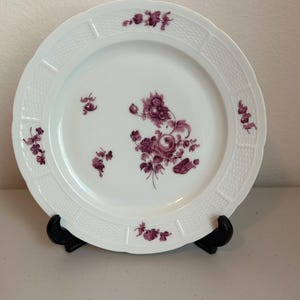 May include: White porcelain plate with a decorative floral design in shades of purple. The plate has a textured rim and is displayed on a black stand. The floral pattern is centered on the plate and also appears around the edges.