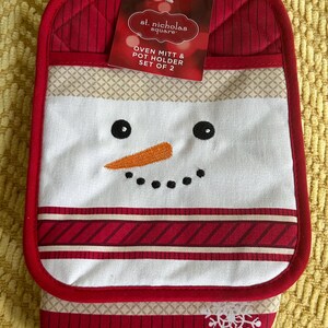 May include: A red and white snowman-themed oven mitt and pot holder set. The mitt features a snowman face with black eyes, a carrot nose, and a stitched mouth. The set includes the text "st. nicholas square" and "OVEN MITT & POT HOLDER SET OF 2".