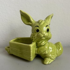 May include: A green ceramic bunny planter with a cart. The bunny has large ears and black eyes. The cart is rectangular with a slatted design. A charming decorative piece for Easter or spring.