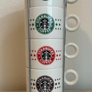 May include: A stack of four white Starbucks coffee mugs with the Starbucks logo in black, green, red, and brown. Each mug has a handle and is held in a silver metal rack. The mugs are decorated with stars.