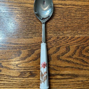 May include: A stainless steel spoon with a white handle featuring a floral design in shades of pink and brown. The spoon is set against a wooden background, showcasing its reflective surface.