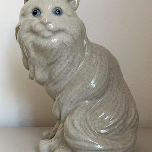 May include: A ceramic cat figurine in a seated position. The cat is off-white with a crackle glaze and blue eyes. The cat has a fluffy appearance with detailed fur texture.