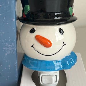 May include: A ceramic snowman night light featuring a black top hat, orange carrot nose, and blue scarf. The snowman has a smiling face and a switch on the bottom. Designed for a UK electrical outlet, it's a festive home decor item.