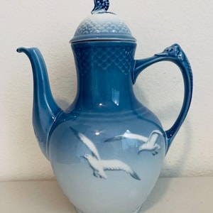 May include: A blue and white ceramic coffee pot with a lid and handle. The pot features a design of seagulls in flight and is a classic piece of kitchenware. The handle has a decorative bird design.