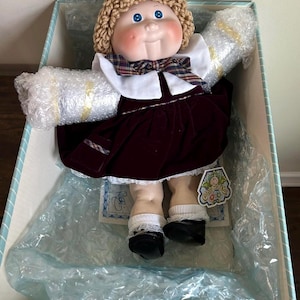 May include: A vintage doll with blue eyes, rosy cheeks, and a crocheted blonde hairstyle. The doll wears a burgundy velvet dress, a white collar with a plaid bow, and black shoes. It is packaged in a blue and white striped box with bubble wrap.