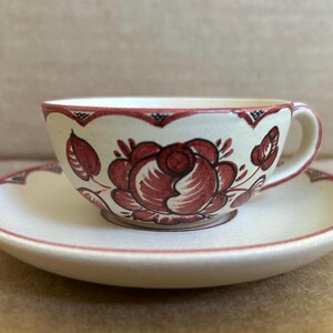 May include: Cream-colored teacup and saucer set with a floral design in shades of red. The cup and saucer have a scalloped edge with a red trim. The teacup features a large rose motif.
