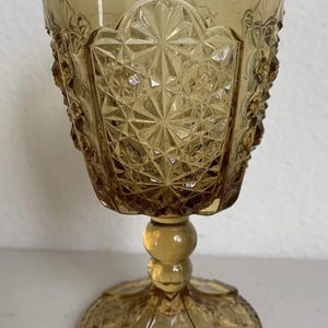 May include: Amber-colored vintage goblet with intricate diamond and floral patterns. The glass features a round base, a bulbous stem, and a detailed bowl. A decorative piece for serving beverages.