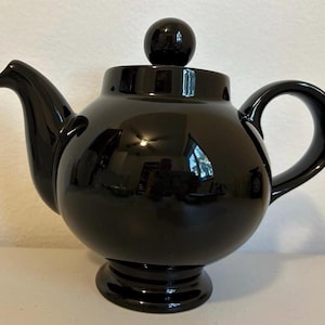 May include: A glossy black teapot with a rounded body, a curved spout, and a loop handle. The lid is topped with a small, round knob. The teapot sits on a pedestal base. The design is simple and elegant.