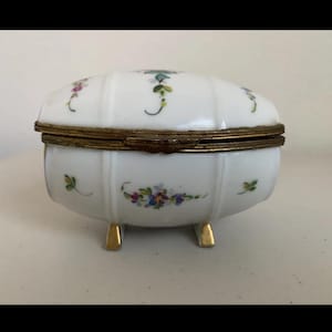 May include: A white porcelain trinket box with a hinged lid and brass trim. The box is decorated with floral designs in shades of blue, purple, and green. It rests on three small gold-colored feet. The box has a rounded shape.