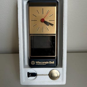 May include: A vintage clock with a gold-colored face, black hands, and a red second hand. The clock is rectangular with a black base and the text "Wisconsin Bell" printed on the front. It includes a gold-colored pendulum.