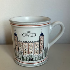 May include: White ceramic mug featuring a detailed illustration of the Tower of London, with the text "THE TOWER" above the image. The mug has a handle and is trimmed with red and gray bands.