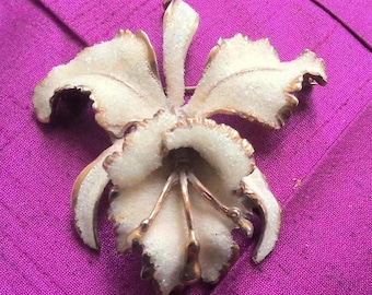 May include: A white orchid brooch with a gold tone finish. The orchid is made of metal and has a textured surface.