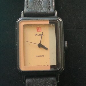May include: A black square-shaped watch with a black leather strap. The watch face is cream-coloured with black hands and a black and white background. The watch face also has the text "ALBA" and "QUARTZ" printed on it.