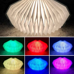 May include: A white paper lamp with a pleated design, shown in six different colors: green, red, white, blue, pink, and off. The lamp is shaped like a clamshell and has a soft, warm glow.
